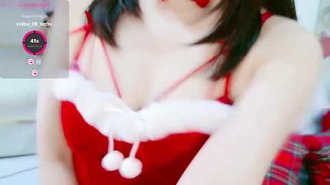 Media: A close-up video of a young woman in a red Santa-themed dress with white fur trim, wearing a red headband. She has a fair complexion and dark hair, partially visible. The background is blurred, focusing on her attire.