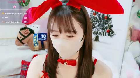 Media: Video of a young Asian woman in a red bunny costume, wearing a face mask, restrained by a black strap around her neck, and standing in a festive, decorated room with a Christmas tree in the background.