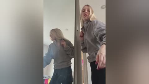 Media: Video of a blonde woman with fair skin, wearing a grey sweatshirt and black pants, leaning against a mirror in a dimly lit room, her reflection blurred.