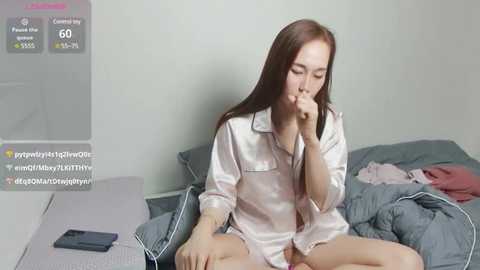 Media: A video of an Asian woman with long brown hair, wearing a white satin pajama top, sitting on a bed with messy gray bedding, holding a cell phone to her ear.