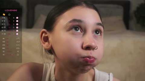 Media: Video of a young, fair-skinned girl with dark hair and a septum piercing, wearing a sleeveless top, in a dimly-lit bedroom with a bed and calendar in the background.