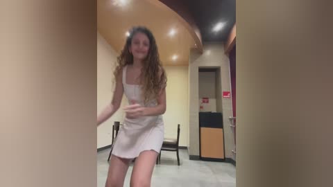 Media: Video of a young woman with long curly hair wearing a white dress, standing in a beige, curved-walled room with recessed lighting and a wooden counter.