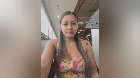 Media: Video of a Latina woman with long, straight, gray-blonde hair, wearing a floral tank top, standing indoors near a window. Background shows an industrial building with exposed beams and a white ceiling.