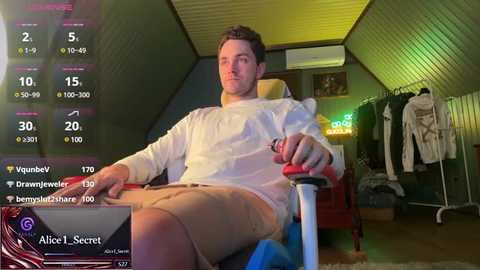 Media: Video of a man sitting in a dimly lit room, wearing a white shirt and beige shorts, holding a controller, with a virtual reality headset and game screen visible.