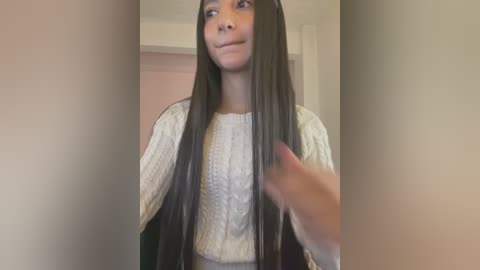 Media: Video of a young woman with long, straight black hair, wearing a cream-colored cable-knit sweater, smiling and holding a section of her hair. Background shows a beige wall and a door, indoors, likely a bedroom.