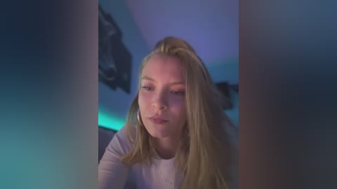 Media: Video of a young woman with long blonde hair, fair skin, and a slight smile, wearing a white top, standing in a dimly lit room with blue and purple lighting.