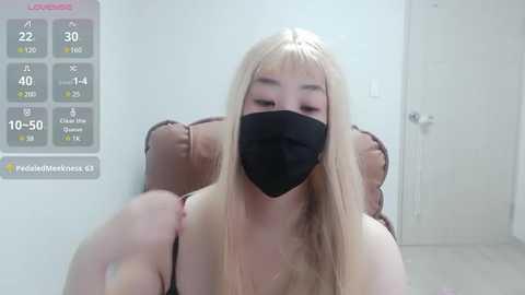 Media: Video of a young Asian woman with long blonde hair, wearing a black face mask, seated in a brown chair. Background includes a white door and a temperature gauge.