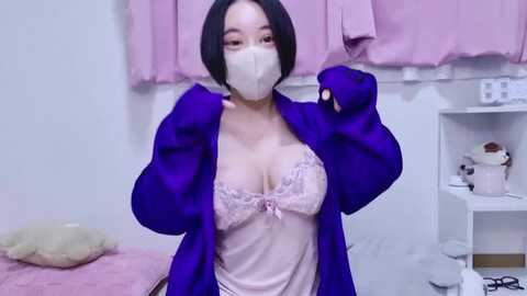 Media: Video of a young Asian woman with short black hair, wearing a pink lace bra and purple robe, a face mask, and gloves, in a pastel-pink room.