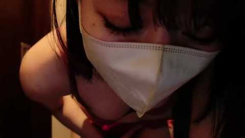 Media: A video captures a close-up of a woman with brown hair wearing a white face mask, eyes closed, and a red bra strap visible. The dim lighting creates a shadowy, intimate atmosphere.