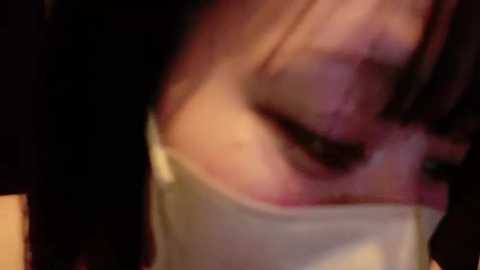 Media: A blurry video of a person wearing a white face mask, with visible skin texture and dark hair, capturing a close-up of the nose and mouth area in dim lighting.