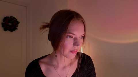 Media: Video of a young Caucasian woman with fair skin, light brown hair in a messy bun, wearing a black top, standing indoors with a Christmas wreath on the wall and a warm, pinkish glow.