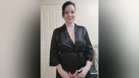 Media: Video of a middle-aged woman with fair skin and dark hair tied back, wearing a black satin robe with a deep V-neck, standing in a hallway with white walls and a closed door.