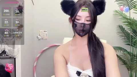 Media: Video of a young woman with long black hair, wearing a black face mask, black cat ears, and a white lace top. Background includes a plant and a pink hula hoop.