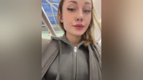 Media: A video of a young Caucasian woman with light skin, blonde hair, and light makeup, wearing a grey hoodie, taken indoors with a blurred background of a large window.