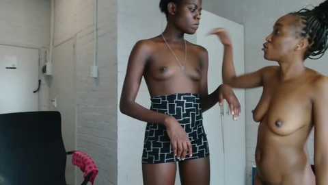 Media: Video of two topless Black women with different body types, one wearing a high-waisted skirt, the other with natural breasts, in a minimalist room with white walls and a black chair.