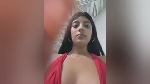 Media: A video of a young, light-skinned woman with long black hair, wearing a low-cut red dress, standing indoors with a geometric-patterned ceiling visible. The image is slightly blurry with a reddish hue.