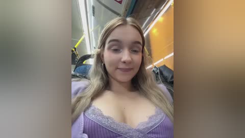 Media: A video of a young woman with long blonde hair, wearing a purple top with lace trim, sitting in a bus. The background shows bus seats and yellow lighting.