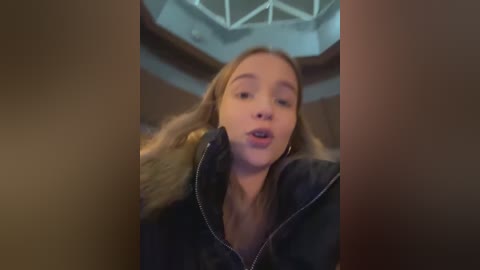 Media: Video of a young Caucasian woman with fair skin and long, wavy blonde hair, wearing a black jacket with fur trim, looking surprised with parted lips, standing indoors with a geometric ceiling in the background.