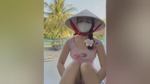 Media: Video of a young woman with light skin, wearing a pink floral dress, white conical hat, and face mask, seated on a poolside deck with palm trees and greenery in the background.