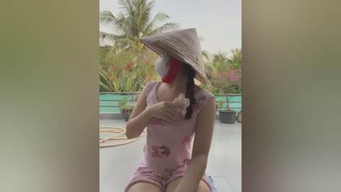 Media: A video of a woman with a conical hat and pink pajamas, holding a pink scrunchie, on a rooftop garden with lush greenery and a turquoise pool in the background.