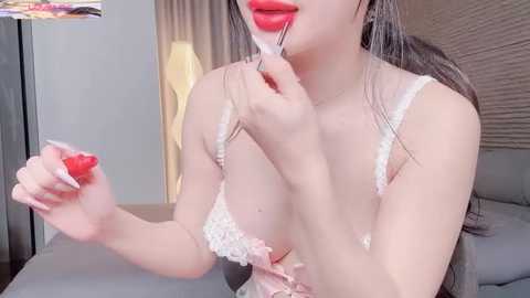 Video of a light-skinned Asian woman with long, straight brown hair, wearing a white lace bra and red lipstick, applying it to her lips. Background features a modern bedroom with beige curtains and a bed.