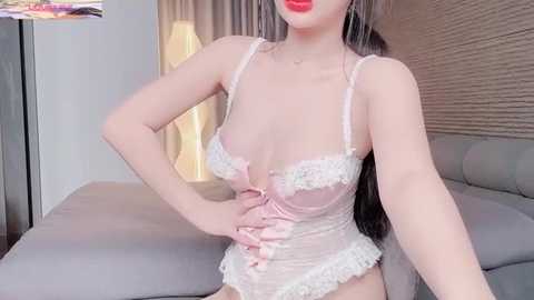 Video of an East Asian woman with long dark hair, wearing a sheer pink lace lingerie bodysuit, sitting on a gray couch in a modern living room with beige walls and a lamp.