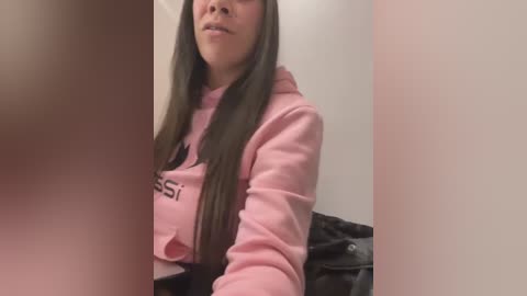 Media: Video of a young woman with long, straight dark hair, wearing a pink hoodie, sitting on a dark sofa against a plain white wall. The image is slightly out of focus.