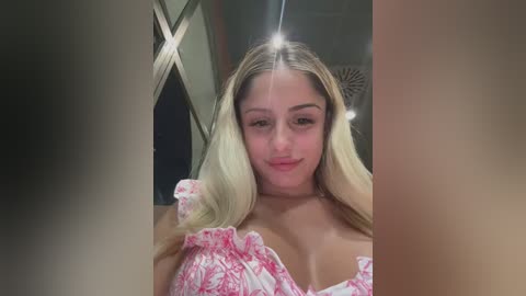 Media: Video of a young woman with long blonde hair, wearing a pink floral dress, standing indoors with a diamond-shaped mirror and decorative objects in the background.