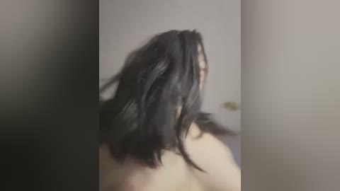 Media: Video of a person with shoulder-length, dark, messy hair, facing away, possibly topless, blurred background, neutral indoor setting.