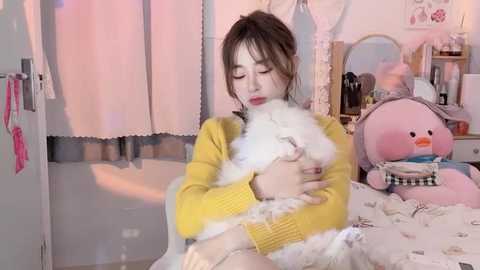 Media: Video of a young Asian woman with light skin, brown hair, and closed eyes, holding a white, fluffy cat in a yellow sweater. Background features pink curtains, a plush Kirby toy, and a white bed with a patterned blanket.