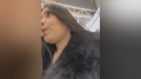 Media: Video of a woman with long, straight dark hair, wearing a black fur coat, blowing a kiss to the left. Background shows an indoor setting with metal beams.