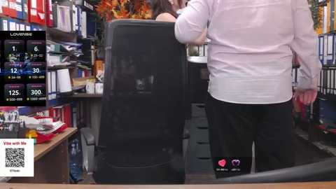 Media: Video of a woman in a white shirt and black pants standing behind a black office chair in a cluttered office. The background features bookshelves, a desk with papers, and a live stream screen displaying a \"VLOG\" watermark.