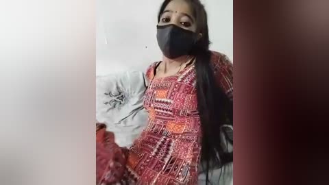 Media: Video of a South Asian woman with long black hair, wearing a red and orange patterned dress, black mask, and seated on a grey couch with floral cushion.