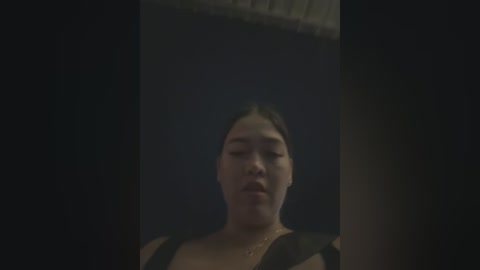 Media: A dimly lit video of an Asian woman with short, dark hair, wearing a black sleeveless top, standing in a dark, narrow hallway.