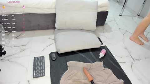 Video of a nude person sitting on a white couch, wearing a beige shirt, holding a dildo, in a modern, white marble-floored room.