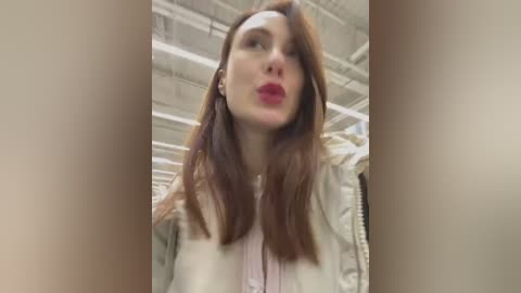 Media: A video of a woman with long, reddish-brown hair and light skin, wearing a beige jacket, red lipstick, and a white headband, taken indoors under fluorescent lighting.