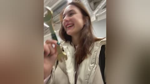 Media: Video of a woman with straight brown hair, wearing a beige puffer jacket, holding a magnifying glass, smiling, in an indoor setting with a white ceiling and soft lighting.