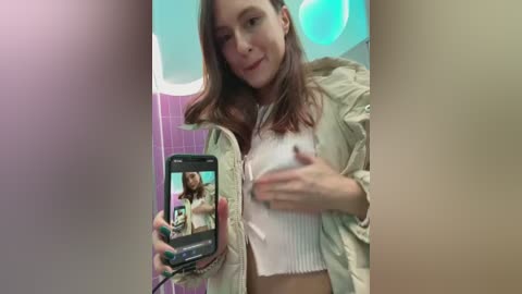 Media: Video of a young Caucasian woman with light skin and long brown hair, wearing a light jacket and a white crop top, taking a selfie in a bathroom with pink tiles and a blue lightbulb.