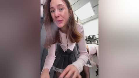 Media: Video of a fair-skinned woman with long brown hair, wearing a pink sweater and black pleated skirt, bending over to pick up a white object in a dimly lit room.