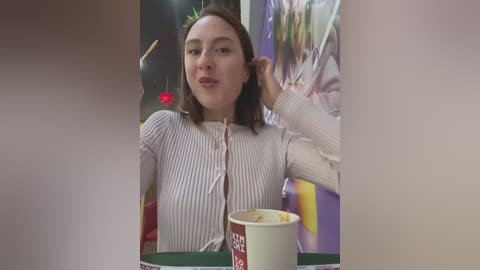 Media: Video of a young woman with straight brown hair, wearing a pink and white striped knit sweater, smiling while drinking coffee at a caf\u00e9 with colorful posters in the background.