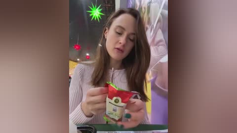 Media: Video of a young woman with light skin and brown hair, wearing a white striped sweater, examining a small green and red gift box in a brightly lit room with colorful decorations.