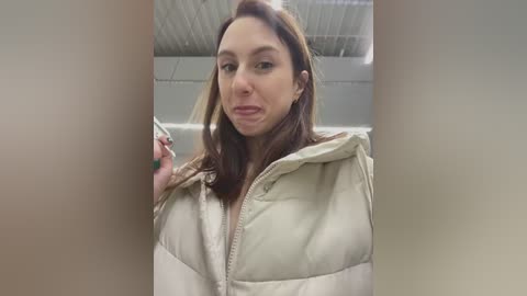 Media: A video of a young Caucasian woman with straight, shoulder-length brown hair, wearing a beige puffy jacket. She is indoors, with a blurred industrial ceiling in the background, and is making a face with her lips pursed.