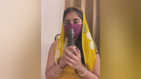 Media: Video of a young South Asian woman wearing a yellow sari with white polka dots, holding a large, black dildo. She has a purple face mask on and is indoors against a beige and brown curtain backdrop.