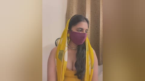 Media: A video of a South Asian woman with long, dark hair, wearing a yellow sari, maroon face mask, gold earrings, and a pearl necklace, indoors against beige and brown curtains.