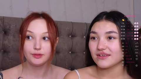 Media: Video of two women with fair skin and long, straight hair (red and black), seated side by side against a tufted gray headboard. They wear light-colored tops.