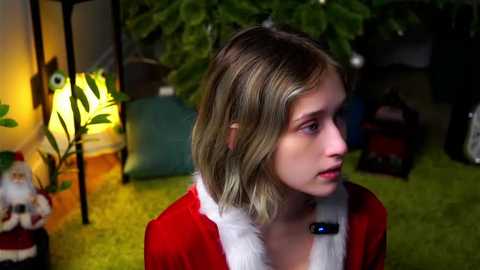 Media: Video of a young Caucasian girl with shoulder-length blonde hair, wearing a red velvet Santa costume with white fur trim, standing indoors next to a lit Christmas tree and festive decorations.