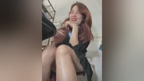 Media: Video of a smiling East Asian woman with long, wavy brown hair, wearing a black robe, sitting on a toilet in a tiled bathroom with a beige wall.