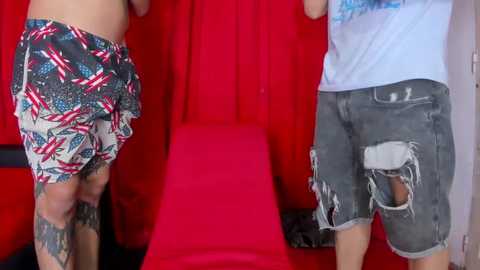 Media: Video of two men facing each other, both wearing distressed denim shorts, standing on a red cushioned bench in front of a red curtain backdrop.