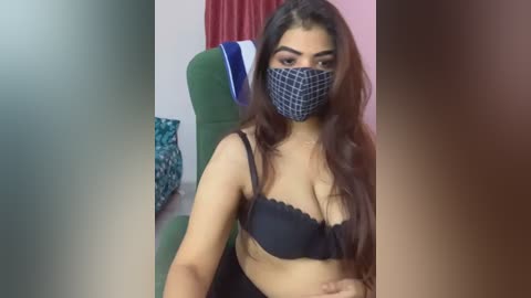 Media: Video of a South Asian woman with long, wavy brown hair and medium skin tone, wearing a black bra and a face mask, sitting on a green chair in a room with a blue and white patterned pillow and red curtains.