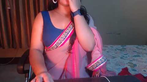 Media: Video of a South Asian woman in a blue and pink sari, seated at a desk, wearing a black watch, in a dimly lit room with floral wallpaper and a bed.